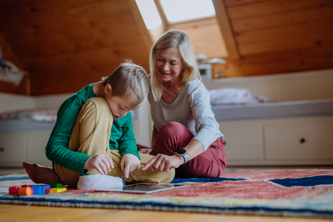 Child with down Syndrome Sitting on Floor and Using Tablet with Grandmother at Home.